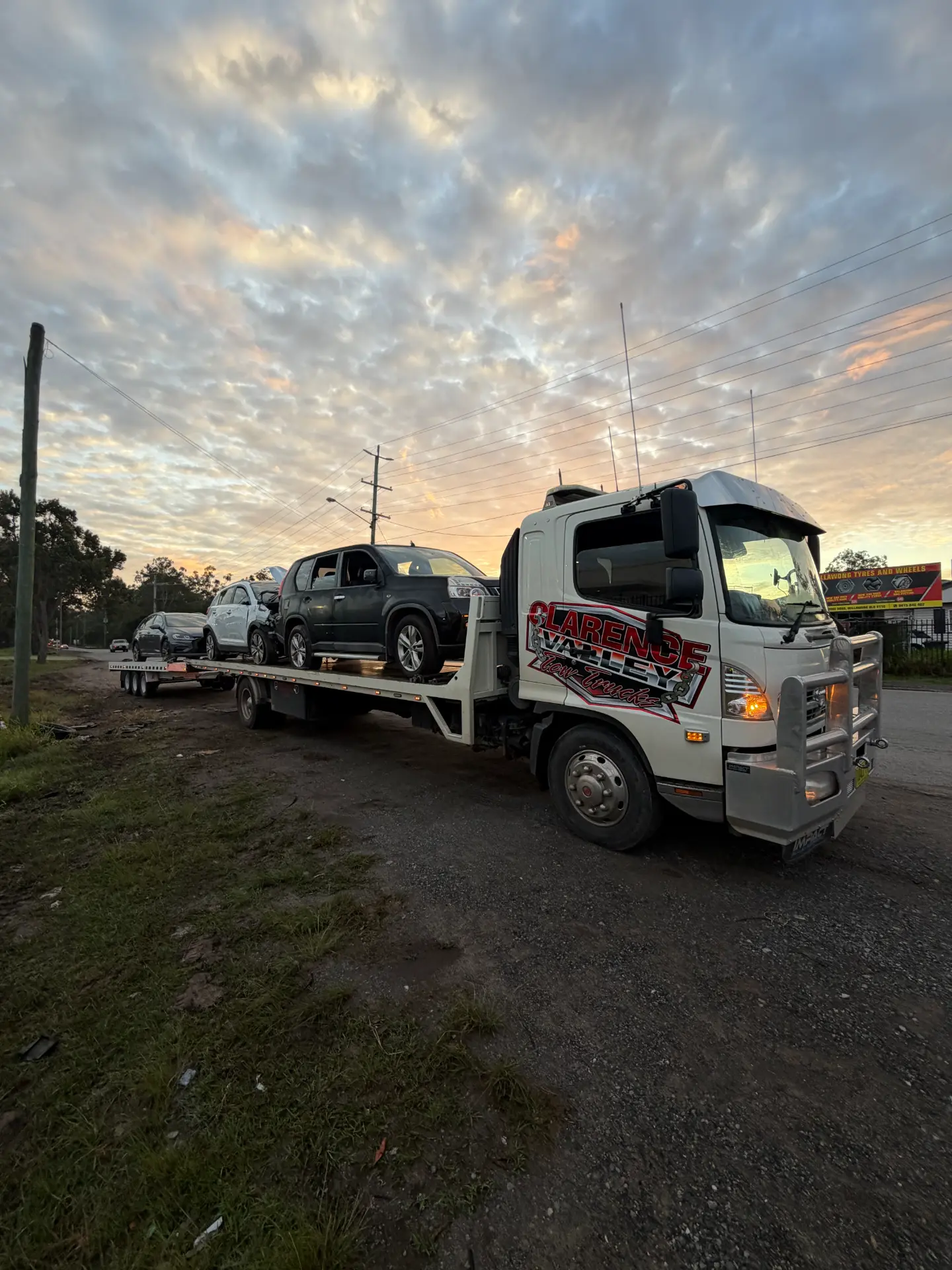 Two crash-damaged cars stacked on the Clarence Valley Tow Trucks tilt tray at dusk