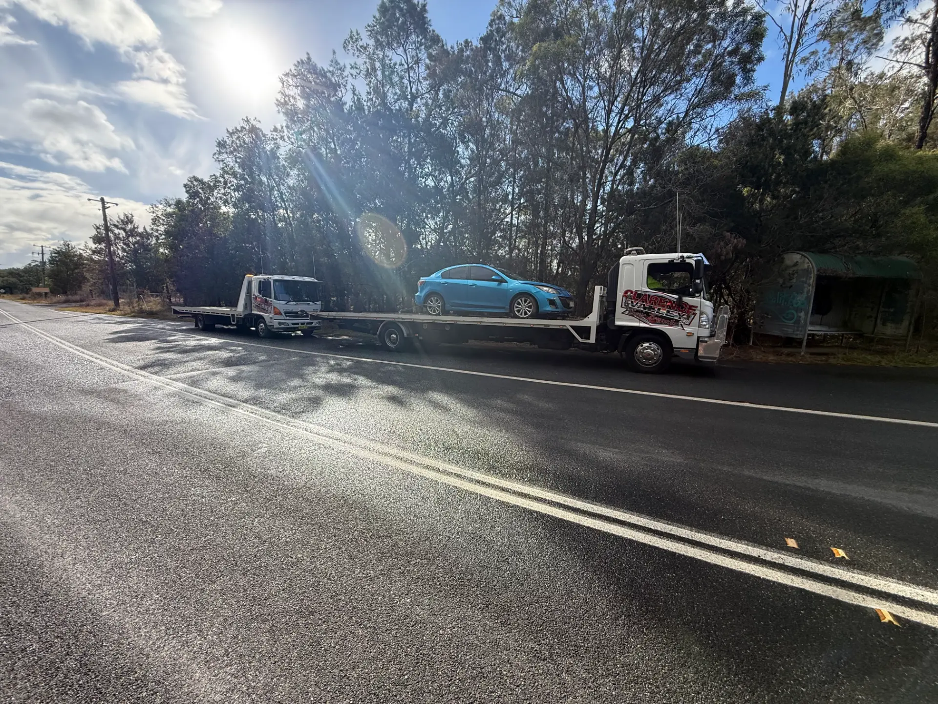 A blue sedan loaded onto the tilt tray beside a country road in low sun