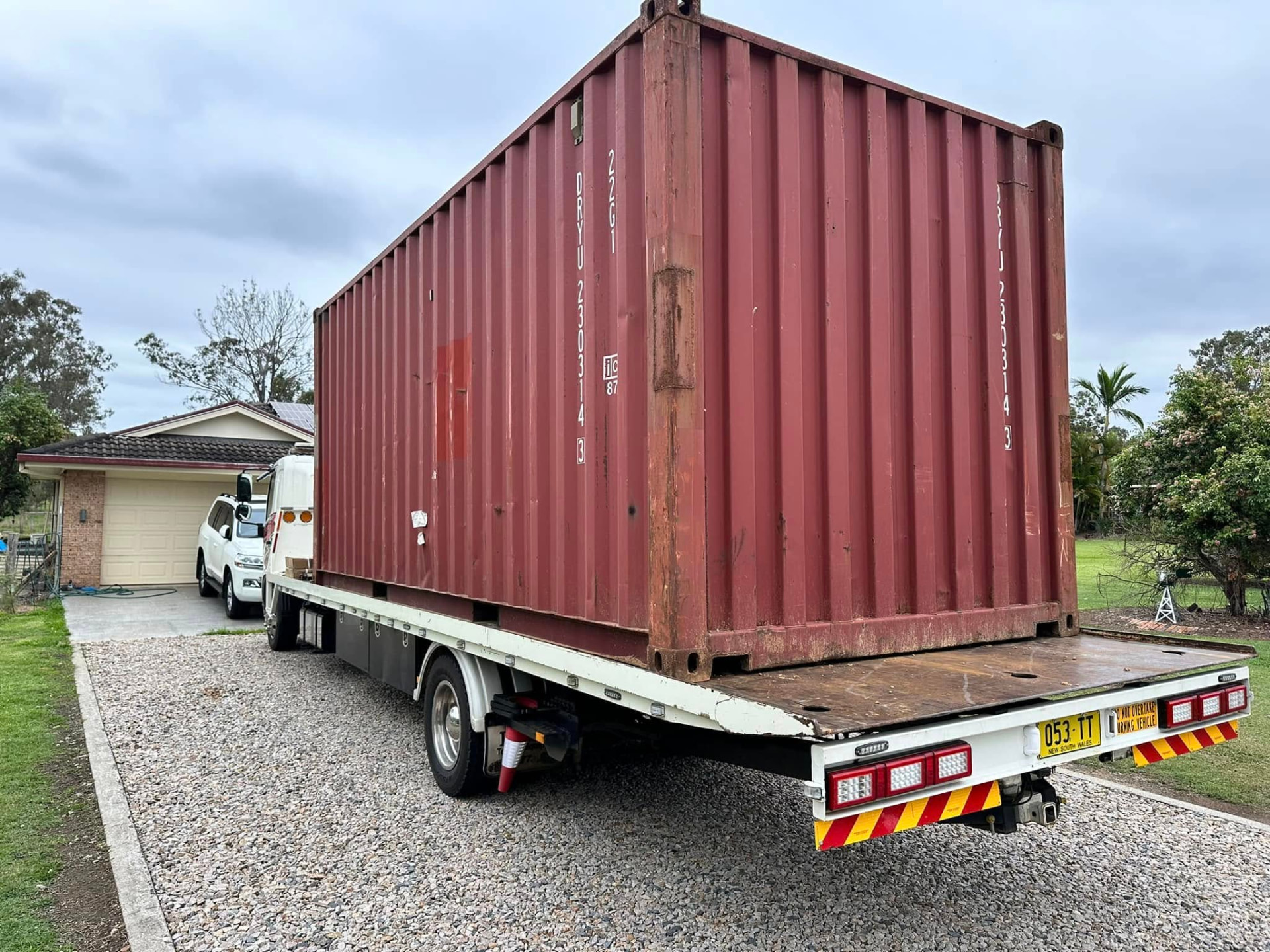 Red shipping container being delivered by tilt tray onto a gravel driveway at a Clarence Valley home