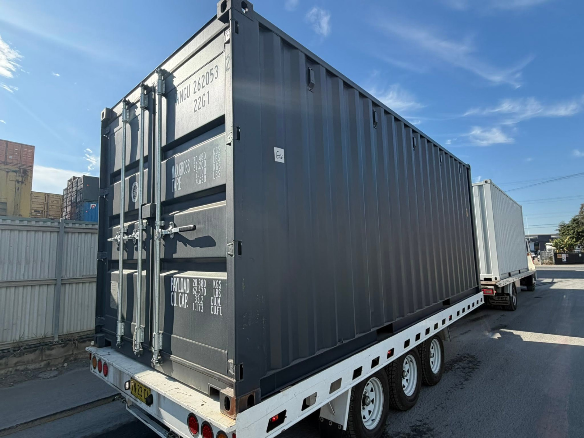 Charcoal 20 foot shipping container strapped to a tilt tray trailer in a container yard